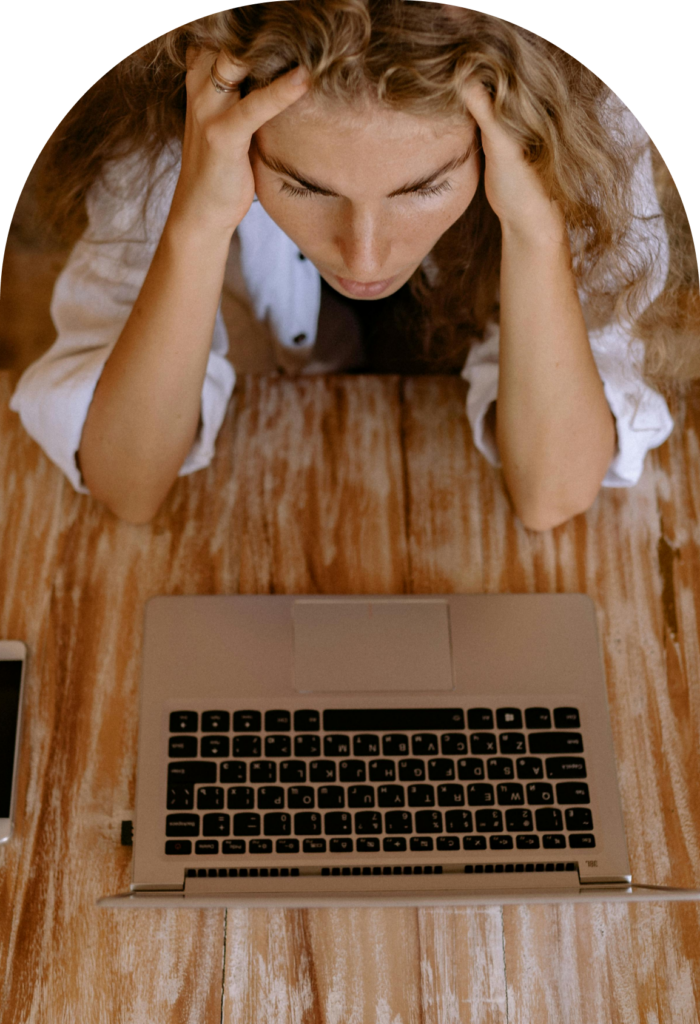 Person sits at a wooden desk, hands in her hair, looking down at a laptop with a smartphone nearby, suggesting focus or stress.