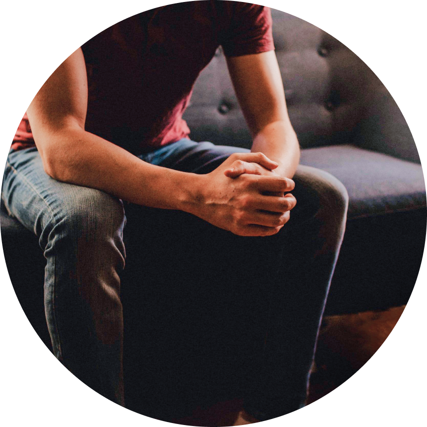 Person sitting on a dark sofa with hands clasped in their lap, wearing a red shirt and jeans.