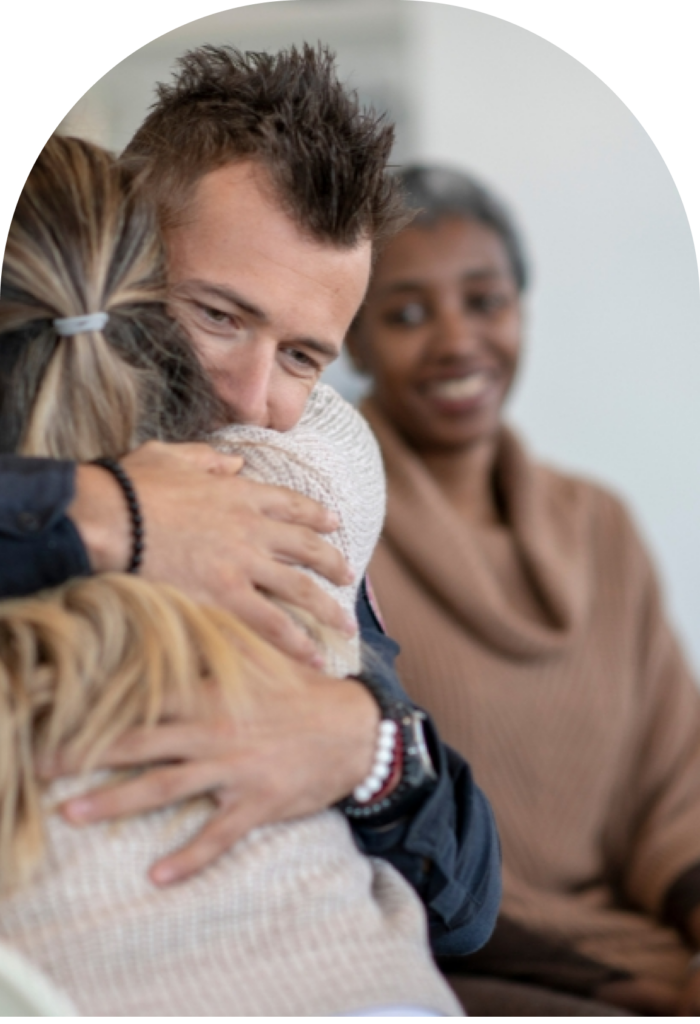 Man hugs a child in a warm embrace as a smiling person watches in the background.