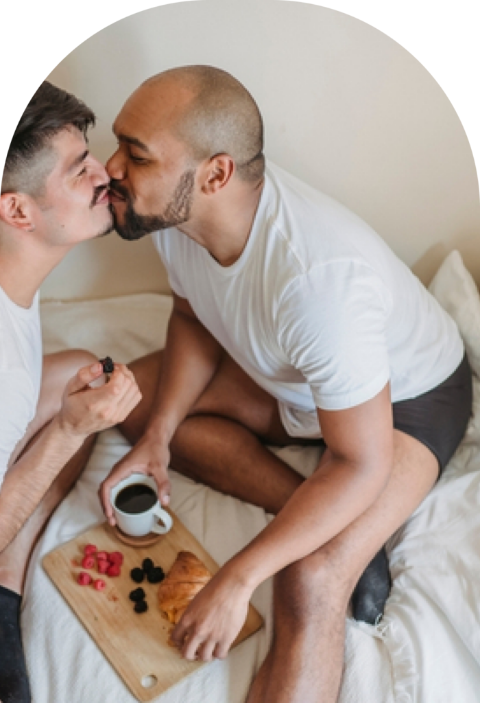 Two men in white shirts kiss while sitting on a bed, sharing a breakfast tray with coffee and pastries nearby.
