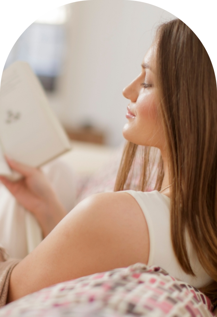 Side profile of a woman with long brown hair resting in bed while reading a book with her eyes closed.