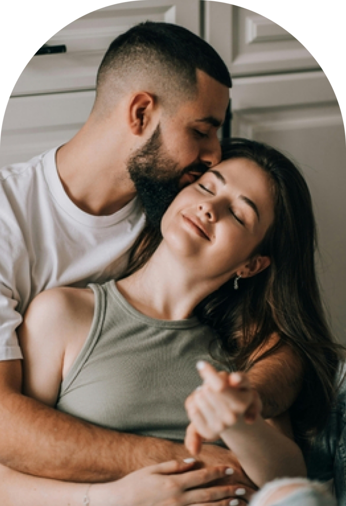 Man kissing a smiling woman on the head as they cuddle indoors.