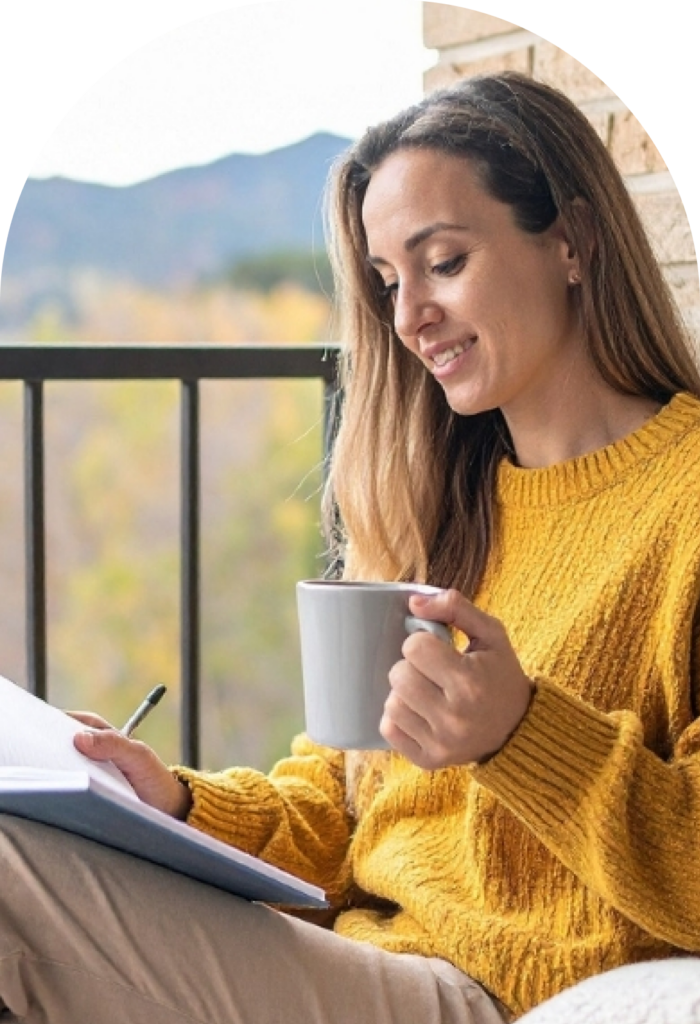 Woman with a mug writing in a notebook on a balcony with mountain view.