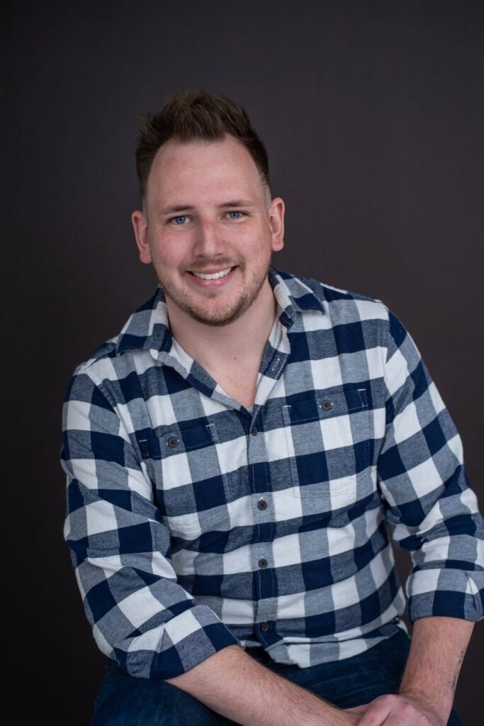 Portrait of a smiling man wearing a blue and white plaid shirt, seated against a dark backdrop.