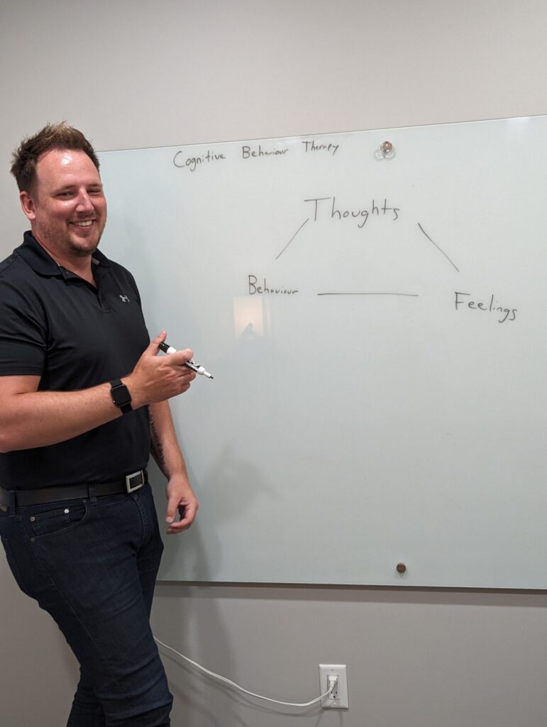 Smiling man in a black polo standing beside a whiteboard with 'Cognitive Behavior Therapy' and a simple diagram about thoughts, behaviour, and feelings written on it.