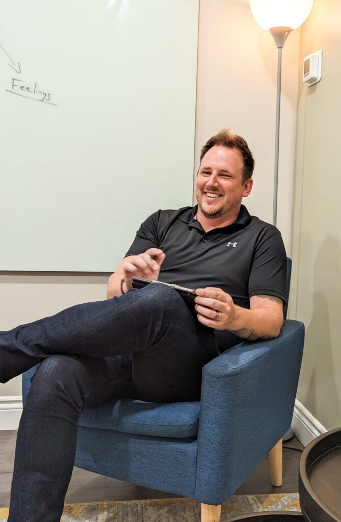 Man in a black polo shirt sits in a blue armchair, smiling, holding a notebook in a bright office setting with a whiteboard nearby.