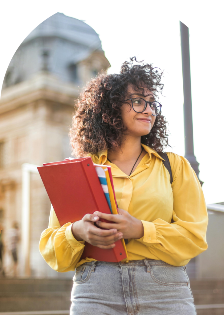 Young woman with curly hair and glasses holding books outside Stone Silo building.